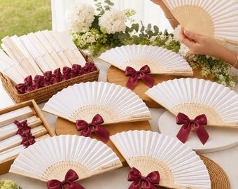 May include: A collection of white folding hand fans with light wood ribs, each featuring a deep burgundy satin ribbon bow at the base. Some fans are displayed open on circular wooden mats, while others are neatly stacked in a woven basket.