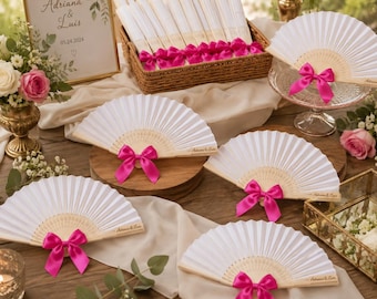 May include: A collection of white folding hand fans with light wood ribs, each adorned with a bright pink satin ribbon bow. The fans feature the text Adriana & Luis printed on the side. A framed sign displays the same names and the date 05.24.2024.