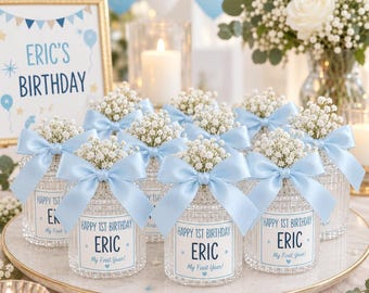 May include: A collection of clear glass jars decorated with light blue satin bows and white baby's breath flowers. Each jar features a label that reads Happy 1st Birthday Eric My First Year, arranged on a round white tray with a birthday sign.