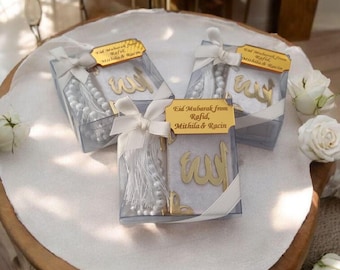 May include: Three clear gift boxes tied with white ribbons, each containing a white prayer bead set, a gold-colored Arabic calligraphy piece, and a small white towel. The boxes have gold tags that read "Eid Mubarak from Rafid, Mithila & Racin."