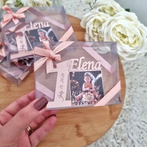 Könnte beinhalten: Transparente Plastikboxen mit rosa Schleife, die ein Babyfoto mit dem Namen "Elena" und dem Datum "18. Juli 2026" enthalten. Die Boxen liegen auf einer Holzoberfläche, im Hintergrund weiße Blumen.