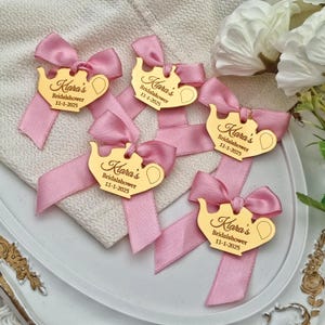 May include: Pink satin ribbon bows with gold-colored teapot-shaped tags. Each tag is engraved with the text "Kiara's Bridalshower 11-1-2025". The bows are arranged on a white surface, suggesting a celebratory event.