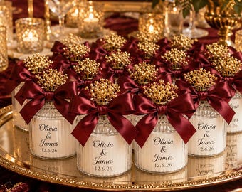 May include: A wedding reception table setting with candles, flowers, and favors. Small glass jars with burgundy bows and gold accents are arranged on a gold tray. A sign reads "Olivia & James 12.6.26".