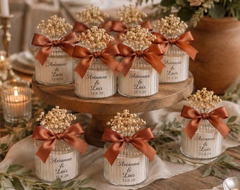 May include: A collection of small, clear, ribbed glass jars filled with white material, each topped with a rust-colored satin ribbon bow and a sprig of dried baby's breath. The jars feature the printed text Arianna & Luis 12.6.26 on the front.
