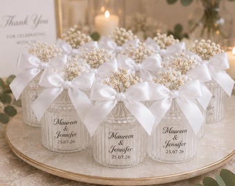 May include: A collection of small, clear, ribbed glass jars, each adorned with a white satin ribbon bow and filled with dried baby's breath flowers. Each jar features the printed text 'Maureen & Jennifer' followed by the date '25.07.26'.