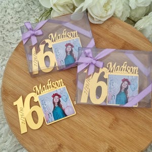 May include: Gold-colored "16" and "Madison" photo holders with a picture of a young woman, presented in clear boxes tied with lavender ribbons. The date "7/25/10" is also visible. The items are arranged on a wooden surface.