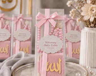 May include: A clear box tied with a pink ribbon contains a pink prayer bead necklace, a small pink book, and a pen. The box has a label that says "Welcoming Baby Zahra 17.01.2026". The book has gold Arabic script.