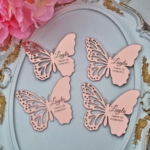 Peut inclure: Quatre décorations en forme de papillon rose. Deux papillons ont des ailes ajourées complexes, tandis que les deux autres ont des ailes pleines. Chaque papillon porte le nom "Layla" et le texte "Sweet 16 02/08/2025". Les papillons sont disposés sur un plateau décoratif.
