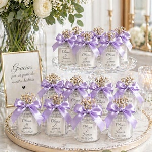 May include: Wedding favors featuring small glass jars with lavender bows and floral accents. Each jar has a label with the names "Olivia & James" and the date "06-21-2026". The favors are arranged on a decorative tray.