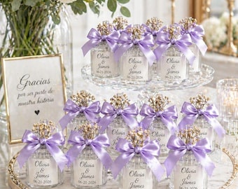 Op de afbeelding: Bruiloftsgunstjes met kleine glazen potjes met lavendelkleurige strikken en bloemaccenten. Elk potje heeft een label met de namen "Olivia & James" en de datum "06-21-2026". De gunstjes zijn gerangschikt op een decoratief dienblad.