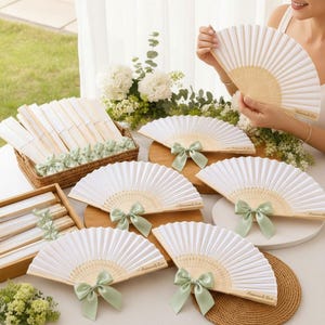May include: A collection of white folding hand fans with light green satin ribbon bows. Each fan features a light wood frame and white fabric. Some fans are displayed open, while others are neatly stacked in a woven basket and wooden tray.