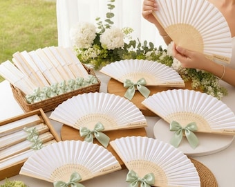 May include: A collection of white folding hand fans with light green satin ribbon bows. Each fan features a light wood frame and white fabric. Some fans are displayed open, while others are neatly stacked in a woven basket and wooden tray.