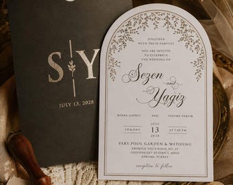 May include: An arched wedding invitation card in white with gold botanical borders and black text for Sezen and Yagiz, paired with a dark charcoal envelope featuring gold foil initials S and Y, and a brass wax seal stamp with a wax seal.