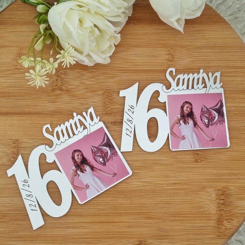 May include: Two white wooden photo holders with the number 16 and the name "Samtya" in script. Each holder has a photo of a young woman in a white dress holding balloons. The date 12/8/26 is printed on the side of the number 16.