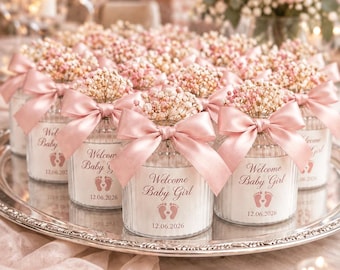 May include: A collection of small glass jars filled with pink baby's breath flowers. Each jar features a pink satin ribbon bow and a label printed with the text Welcome Baby Girl, a pair of baby footprints, and the date 12.06.2026.