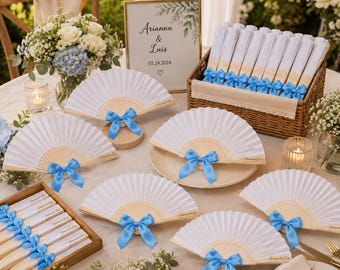 May include: White folding hand fans with light wood ribs and bright blue satin bows. Several fans are displayed open on a table, while others are neatly stacked in a wooden crate. A sign reads Arianna & Luis 05.24.2024 with a heart symbol.