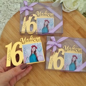 May include: Gold-colored "16" and "Madison" photo frame favors with a picture of a young woman in a teal dress. The frames are in clear boxes tied with lavender ribbons. The date "7/25/10" is engraved on the frame.