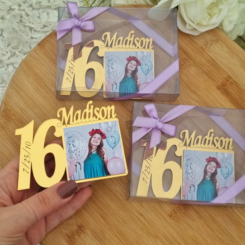 May include: Gold-colored "16" and "Madison" photo frame favors with a picture of a young woman in a teal dress. The frames are in clear boxes tied with lavender ribbons. The date "7/25/10" is engraved on the frame.
