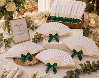 May include: A collection of white folding hand fans with light wood ribs, each adorned with a dark green satin ribbon bow. The fans feature the text Ariamá & Luis printed on the side. A framed sign displays the same names and the date 05.24.2024.