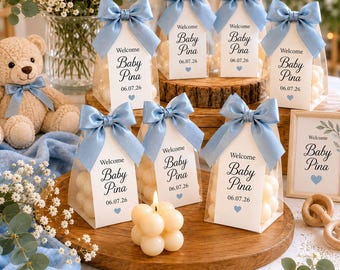 May include: A collection of baby shower favors. Clear boxes filled with round, white candies are adorned with light blue bows and tags that read "Welcome Baby Pina 06.07.26". A teddy bear and a candle are also present.