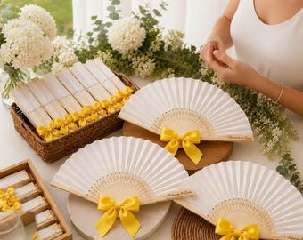 May include: A collection of white folding hand fans with light wood ribs, each adorned with a bright yellow satin ribbon bow. Several fans are displayed open, while others are neatly stacked in a woven basket and a wooden box.