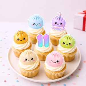 May include: A collection of six cupcakes topped with colourful, smiling dumpling-shaped decorations and a central number 11 topper. The dumplings are blue, yellow, purple with an HBD sign, green, beige, and pink. The number 11 has a gradient finish.