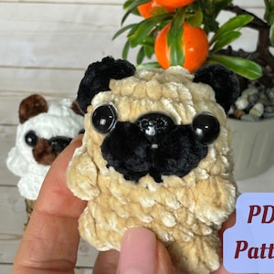 May include: A close-up of two handmade crochet pug dog toys. The front pug is tan with black ears and muzzle, and large black eyes. The second pug is white with brown ears and eyes. The image includes a small plant and the text "PDF Pattern".