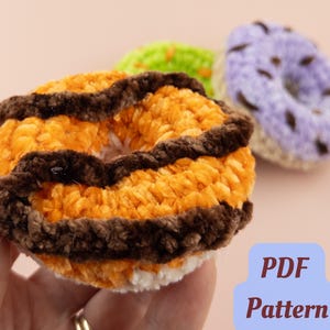 May include: Close-up of a crocheted donut in orange and brown, held in a hand. Two other crocheted donuts in green and purple are in the background. The image includes the text "PDF Pattern".