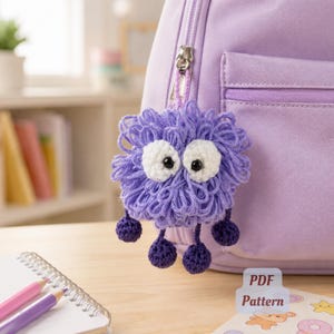 May include: A handmade, purple crochet monster charm with white eyes and black pupils, attached to a lavender backpack. The charm has dangling purple pom-poms. A notebook and pencils are in the foreground. A tag reads "PDF Pattern."