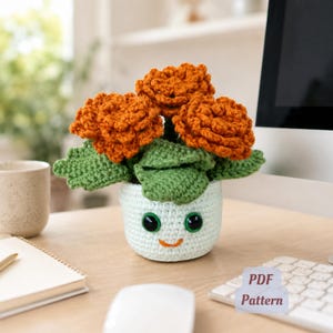 May include: A crocheted flower arrangement in a white pot with a smiling face. The arrangement features orange flowers and green leaves. The pot is on a wooden surface, with a computer monitor and keyboard in the background. The image includes a "PDF Pattern" label.