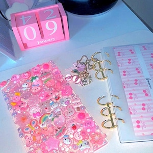 May include: A pink desk setup featuring a bedazzled phone case, a pink wooden calendar displaying January 29, and a clear binder with gold rings. The phone case is covered in various pink charms. A phone sits on a black charging base.
