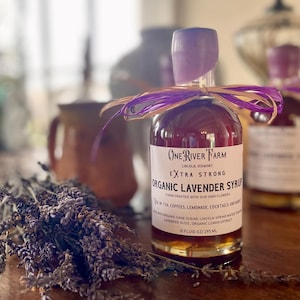 May include: A bottle of One River Farm Organic Lavender Syrup with a purple cap and ribbon. The label reads "Extra Strong" and lists ingredients. A bundle of dried lavender sits nearby. The bottle contains 10 fluid oz / 295 mL.