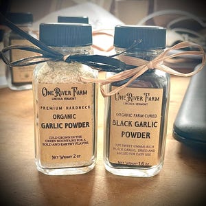 May include: Two glass jars of garlic powder. One jar contains organic garlic powder, the other black garlic powder. Both jars have black lids and are tied with raffia. The labels read "One River Farm" and include the net weight of each jar.