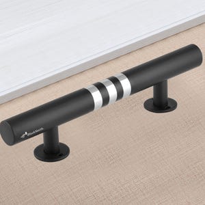 以下が含まれることがあります： 3本のシルバーのストライプが入った黒いドアプル。円筒形のハンドルはマット仕上げで、2つの円形のベースに取り付けられています。「BlackSmith」の文字がハンドルに印刷されています。