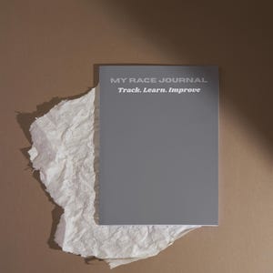 Peut inclure: Un journal gris intitulé "MY RACE JOURNAL" avec le texte "Track. Learn. Improve." Le journal repose sur un morceau de papier blanc froissé sur un fond marron. La couverture du journal est grise unie et mate.