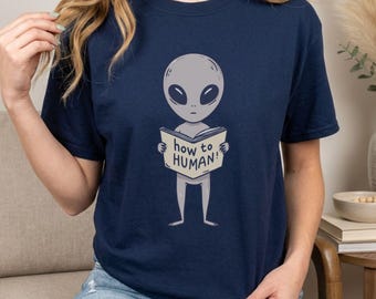 May include: A navy blue short-sleeve t-shirt featuring a graphic of a gray alien holding an open book with the text "how to HUMAN!" printed on the cover. The alien has a large head, dark oval eyes, and a slender body.