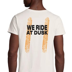 以下が含まれることがあります： 黒とオレンジ色の「WE RIDE AT DUSK」の文字が入った、クリーム色のTシャツ。デザインには、2本のオレンジ色のタイヤ痕が垂直に入っています。このシャツは柔らかく快適な素材で作られています。