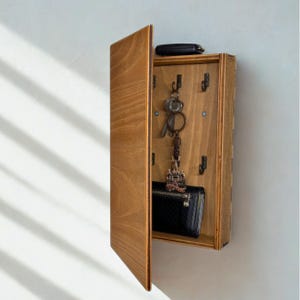 Può includere: Un portachiavi in legno con anta a cerniera, montato su una parete bianca. L'interno presenta ganci per chiavi, un portafoglio nero e una chiave dell'auto. Il legno ha una finitura calda e naturale.