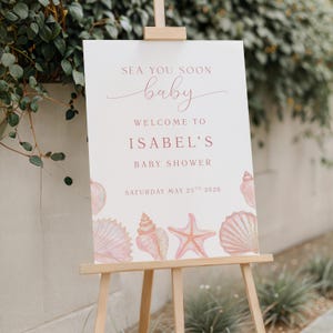 Może przedstawiać: Biały znak na drewnianym sztaludze z napisem "Sea You Soon baby Welcome to Isabel's Baby Shower Saturday May 25th 2026". Znak jest ozdobiony różowymi muszlami i rozgwiazdą.