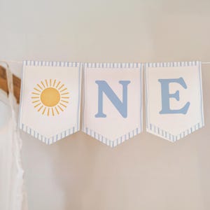 Könnte beinhalten: Ein dekoratives Banner bestehend aus drei an einer Schnur hängenden Wimpeln. Der erste Wimpel zeigt eine gelbe Sonnengrafik, während der zweite und dritte Wimpel die blauen Buchstaben O, N und E tragen. Jeder Wimpel hat hellblaue Streifen.