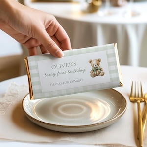 Könnte beinhalten: Ein Schokoriegel mit einer hellgrünen und weißen Vichy-Musterverpackung. Die Verpackung zeigt den Text "OLIVER'S beary first birthday" und eine Teddybär-Illustration. Die Worte "THANKS FOR COMING!" sind ebenfalls aufgedruckt.