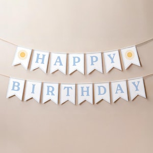 May include: A two-row banner spelling HAPPY BIRTHDAY with light blue letters on white pennants. Each pennant has a thin striped border. The first and last pennants on the top row feature a yellow sun illustration. The banner hangs on a thin string.