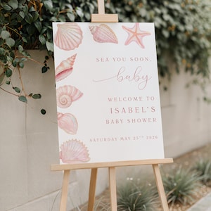 May include: A white sign on a wooden easel featuring pink seashell illustrations. The sign says "Sea You Soon, baby. Welcome to Isabel's Baby Shower" and the date "Saturday May 25th 2026".
