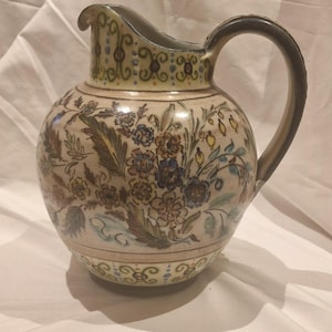 Puede incluir: Jarra de cerámica con diseño floral. La jarra tiene una base de color crema con un patrón de flores y hojas azules, marrones y verdes. El asa y el borde son de color marrón oscuro. Es un objeto decorativo.