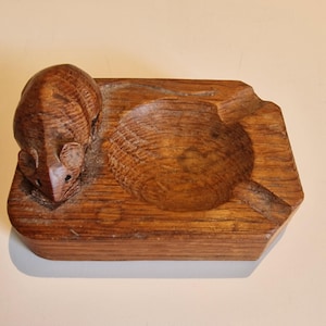 May include: A rectangular brown wooden ashtray featuring a carved mouse figure on one side. The piece has a circular central basin with a smooth, polished finish and visible wood grain patterns throughout the entire carved structure.