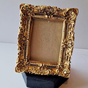 May include: An antique-style, gold-coloured picture frame with elaborate carvings. The frame is rectangular and rests on a small, black, geometric stand. The frame's design includes floral and scroll motifs, typical of the period.