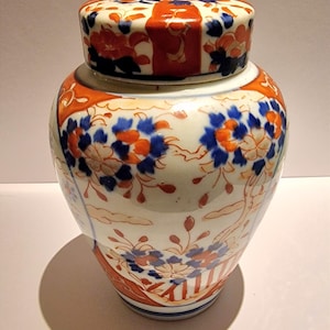 May include: A ceramic ginger jar with a lid, featuring a floral design in orange and blue against a white background. The jar has a rounded body and a matching lid, showcasing a traditional Asian aesthetic.