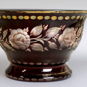 May include: A dark burgundy glass bowl with a wide rim, featuring a floral design of roses and leaves. The rim is accented with gold and green details. The bowl has a pedestal base and is likely for decorative use.