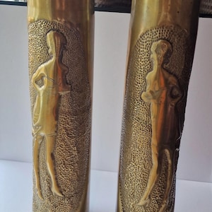 May include: Two antique brass vases with embossed Art Deco style figures. The vases have a textured background and a golden hue. The figures are in a standing pose, adding a touch of vintage elegance.