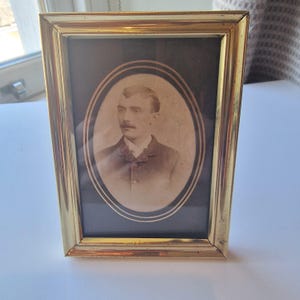 May include: A vintage gold-toned rectangular picture frame with an oval portrait of a man in a dark suit and bow tie. The portrait is in sepia tones, set against a dark background.